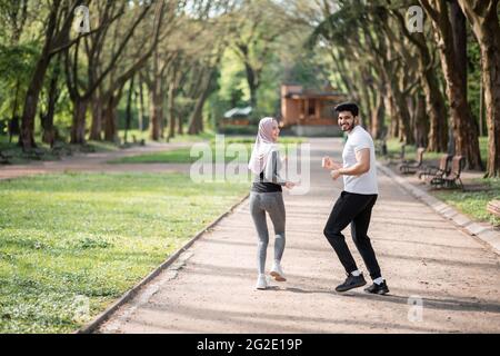 Glückliches arabisches Paar in aktiver Kleidung Joggen im grünen Park während der Morgenzeit. Junge Männer und Frauen genießen die gemeinsame Zeit. Gemeinsames Training im Freien. Stockfoto