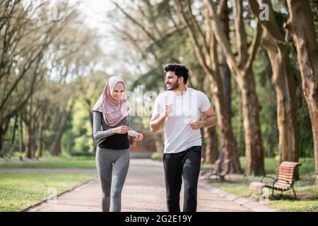 Positive junge Menschen in Sportkleidung laufen zusammen im morgendlichen grünen Park. Gutaussehender muslimischer Ehemann und charmante Frau in Hijab Training aktiv im Freien. Stockfoto