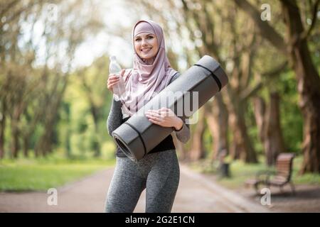 Sportliche, gesunde Frau im Hijab, die Yogamatte und Flaschenwasser hält, während sie im Stadtpark steht. Lächelnde muslimische Frau, die während des Trainings im Freien Pause nimmt. Stockfoto