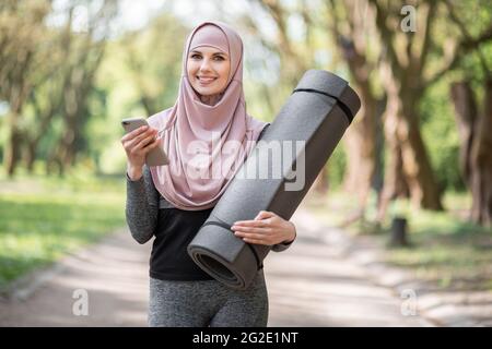 Vorderansicht einer lächelnden Frau im Hijab- und Sportoutfit mit einem modernen Mobiltelefon, während sie beim Training im Freien Pause einnahm. Aktives Lifestyle-Konzept. Stockfoto