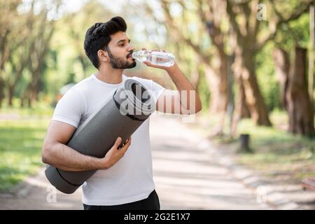 Seitenansicht eines arabischen bärtigen Mannes in Sportkleidung, der frisches Wasser aus der Flasche trank, während er im grünen Park stand. Schöner Athlet, der beim Workout im Freien eine Pause einnimmt. Stockfoto