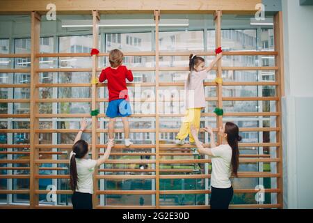 Kinder an der schwedischen Wand üben im Fitnessstudio im Kindergarten oder in der Grundschule mit Lehrern. Sport- und Fitnesskonzept für Kinder Stockfoto