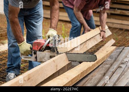 Nahaufnahme von Händen unbekannter Männer, die Kettensäge im Lager oder auf der Baustelle sägen und Schnittholz sägen Stockfoto