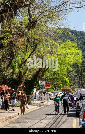 Menschen, die auf einer der Straßen des Higino da Silveira-Platzes in der Nähe einiger hoher Bäume und Autos unter einem sonnigen, klaren, blauen Himmel spazieren gehen. Stockfoto