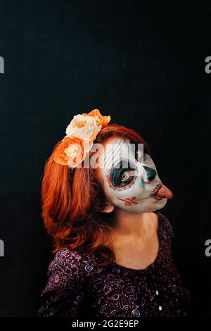 Eine junge Frau am Tag der Toten Maske Schädel Gesicht Kunst. Frau mit Schädel Make-up und roten Haaren zeigt Zunge auf dunklem Hintergrund, Halloween oder Horror-Thema Stockfoto