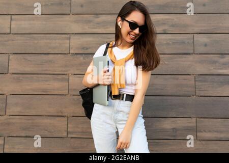 Faszinierend schön lächelnd charmante junge Brunet Mädchen schaut nach unten halten Computer Laptop und schwarze Sonnenbrille in weißem T-Shirt und hellblau Stockfoto