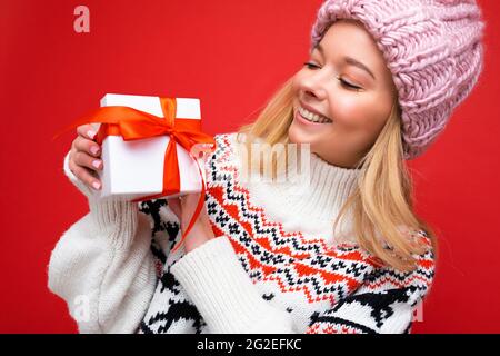 Nahaufnahme einer schönen, fröhlich lächelnden jungen blonden Frau, die über einer roten Hintergrundwand isoliert ist und einen Winterpullover und einen pinken Hut mit weißem Hintergrund trägt Stockfoto