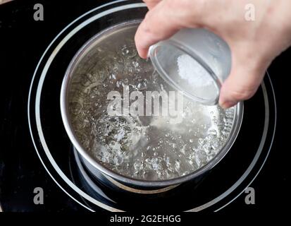 Auf dem elektrischen Induktionskochfeld wird Salz in den kochenden Wassertopf gegeben Stockfoto