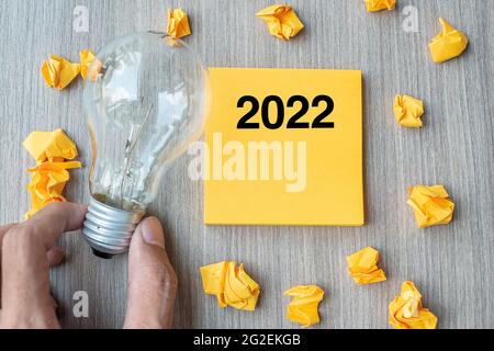 2022 Wörter auf gelber Note und zerbröckeltem Papier mit Geschäftsmann, der Glühbirne auf Holztischhintergrund hielt. Neues Jahr Neue Idee Kreativ, Innovation, Im Stockfoto