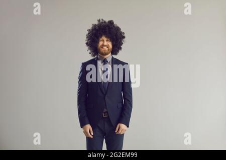 Glücklicher Geschäftsmann in elegantem Anzug und verrückter lockiger Perücke, die im Studio steht und lächelt Stockfoto