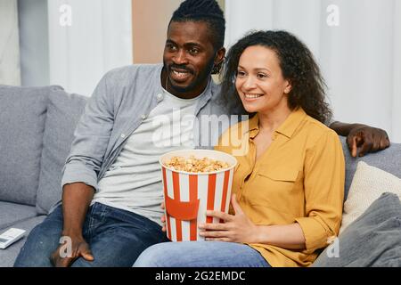 Gemischtes Rennpaar schaut zusammen mit einem großen Eimer Popcorn in ihrem Zuhause fern, während es auf einer Couch sitzt Stockfoto