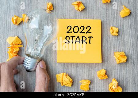 2022 Startworte auf gelber Note und zerbröckeltem Papier mit Geschäftsmann, der Glühbirne auf Holztischhintergrund hält. Neues Jahr Neue Idee Kreativ, Innovati Stockfoto