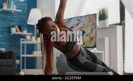 Die schwarze Sportlerin trainiert auf der Yoga-Karte im Wohnzimmer, steht auf der Seitenwand und schaut sich mit einem Laptop ein Fitnessvideo an. Erwachsene in Sportbekleidung Streching Muskel während Strech Gymnastik Stockfoto