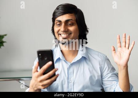 Moderner fröhlicher indischer Kollege mit Smartphone und winkender Hand, fröhlicher hindu-Kerl im formellen Hemd, der den Online-Gesprächspartner begrüßt und die mobile App für virtuelle Videobesprechung verwendet Stockfoto