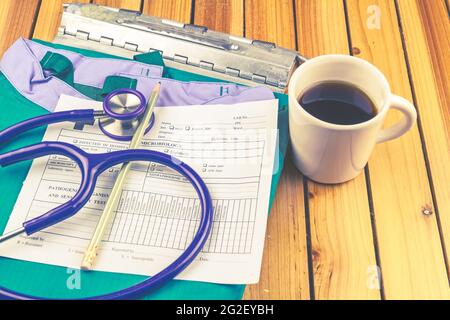 Ein Stethoskop, blaue medizinische Uniform, medizinische Geschichte Ordner Zwischenablage, Bleistift, weiß leer mit Ergebnissen in Spalten und Kaffee Tasse Stapel auf Holz b Stockfoto