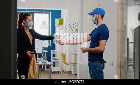 Delivery guy mit Schutz medizinische Gesichtsmaske und Handschuhe gegen Coronavirus bringt Essen zum Mitnehmen Mittagessen Bestellung im Büro des Unternehmens. Geschäftsfrau, die mit einer Smartwatch über kontaktlose POS-Systeme eine Mahlzeit bezahlt Stockfoto