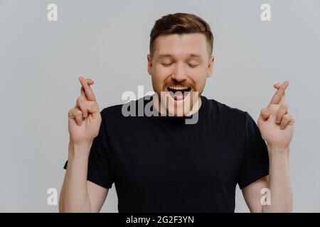Hübscher junger aufgeregt Mann in schwarzem T-Shirt, die Finger kreuzt und auf Glück hofft Stockfoto