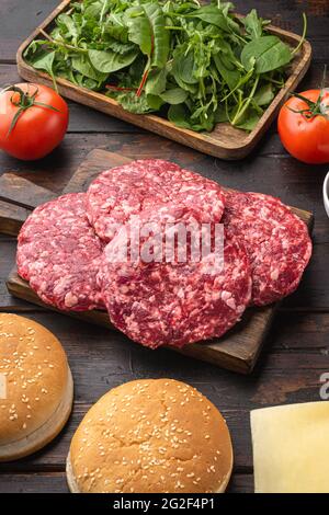 Hausgemachter Hamburger. Rohe Rindersteaks, Sesambrötchen mit anderen Zutaten auf altem dunklen Holztisch Stockfoto