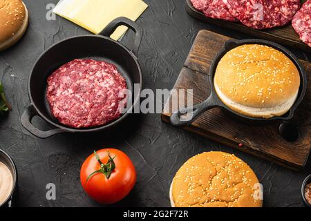 Zutaten zum Kochen von Burgern. Hacksteaks, Brötchen, Tomaten, Kräuter und Gewürze auf schwarzem Stein Stockfoto