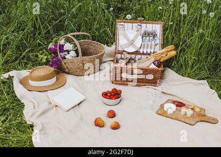 Picknick-Set mit Obst, Käse, Baguette, Erdbeere, Wein mit Blumen in einem Korbkorb auf Plaid. Stockfoto