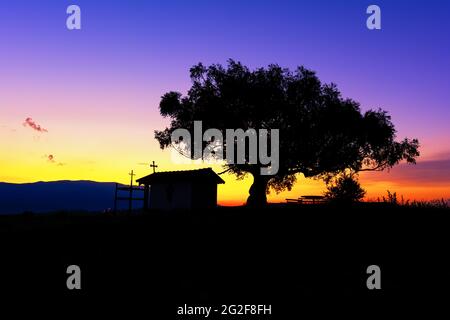 Silhouette der christlichen Kapelle in der Nähe des Dorfes Plana, Bulgarien und ein Baum mit einer großen Krone bei einem malerischen Sonnenuntergang. Stockfoto