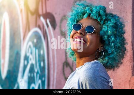 Porträt einer jungen, lächelnden, schwarzen afroamerikanischen Frau mit blauen Haaren und Sonnenbrillen im Freien Stockfoto