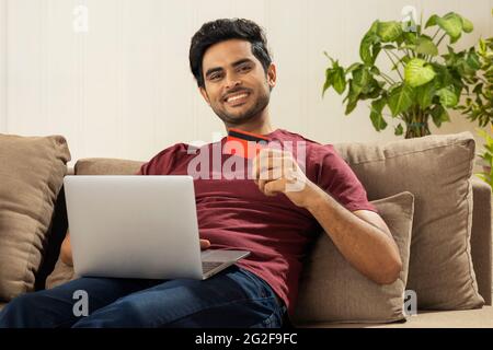 EIN GLÜCKLICHER JUNGER MANN, DER ZU HAUSE SITZT UND ONLINE-TRANSAKTIONEN MACHT Stockfoto