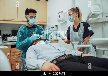 Kaukasischer männlicher Klient liegt auf dem Bett der Ärzte, während männliche und weibliche Zahnärzte auf dem Mund operieren Stockfoto