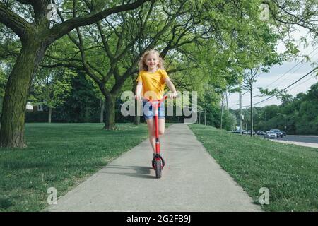 Nette lustige kaukasische Mädchen Kind in gelben T-Shirt Reiten roten Roller auf Straße Road Park im Freien. Sommerspaß umweltfreundliche Sportaktivitäten für Kinder Chil Stockfoto