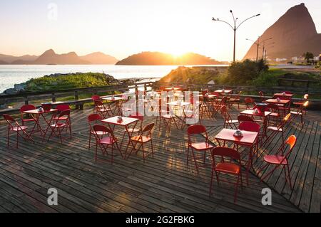 Holzterrasse mit roten Tischen und Stühlen mit Blick auf den Sonnenuntergang und den Zuckerhut - Cafeteria im Freien ohne Personen Stockfoto