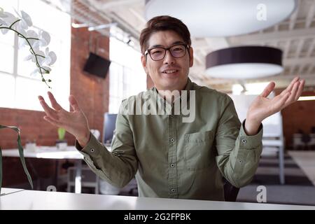 Glücklicher asiatischer Geschäftsmann, der am Schreibtisch sitzt und während eines Videoanrufs redet und gestikuliert Stockfoto