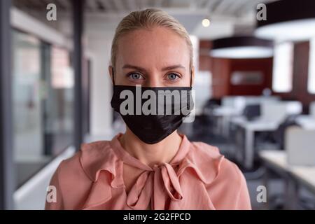 Porträt einer kaukasischen Geschäftsfrau mit Gesichtsmaske, die im Büro steht und vor die Kamera schaut Stockfoto