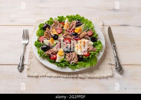 Leckerer Thunfischsalat auf weißem Teller, Messer und Gabel über weißem Holztisch. Verzehrfertig mediterraner Thunfisch-Salat mit Wetheggs, frischem Salat und Tomaten. Stockfoto