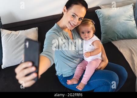 Kaukasische Mutter lächelt, während sie ein Selfie mit ihrem Baby auf der Couch zu Hause sitzt Stockfoto