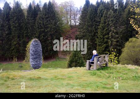 Frau sitzt auf einer Holzbank und bewundert die steinerne 'Fir Cone'-Skulptur The Himalayan Garden & Sculpture Park, Grewelthorpe, Ripon, North Yorkshire. Stockfoto