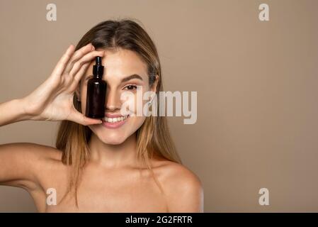 Lächelnde Frau, die Vitamin C-Serum auf beigem Hintergrund in der Nähe ihres Gesichts hält. Hautpflege und Gesundheitskonzept. Stockfoto