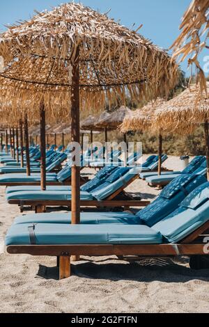 Eine Reihe von leeren blauen Sonnenliegen und Liegen unter den Sonnenschirmen an einem Strand in Mykonos, Griechenland. Stockfoto