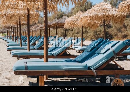 Seitenansicht einer Reihe von leeren blauen Sonnenliegen und Liegen unter den Sonnenschirmen an einem Strand in Mykonos, Griechenland. Stockfoto