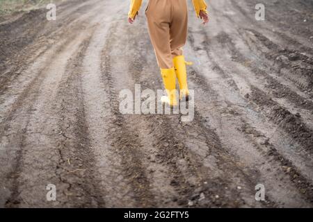 Beine einer Bäuerin in gelben Gummistiefeln, die nach dem Regen durch das kultivierte landwirtschaftliche Feld spazierend. Stockfoto