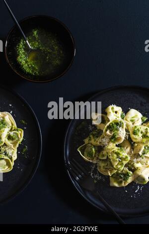 Zwei schwarze Teller auf dunklem Grund mit frischer Tortellini-Pasta mit Pesto-Sauce und einer kleinen schwarzen Schüssel mit hausgemachtem Pesto. Stockfoto