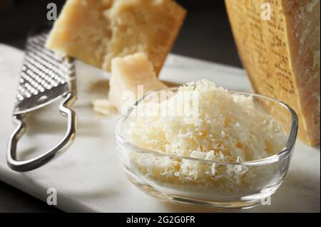 Parmesan in Stücken und gerieben. Glasschale mit geriebenem Parmesan-Käse und Reibe auf Marmorschneidebrett. Nahaufnahme, selektiver Fokus. Stockfoto