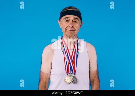 Älterer Athlet mit Sonnenflecken an den Armen, mit drei Medaillen am Hals für eine sportliche Leistung auf blauem Hintergrund Stockfoto