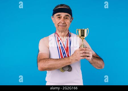 Älterer Athlet mit Sonnenflecken an den Armen, drei Medaillen am Hals und einer Trophäe in den Händen auf blauem Hintergrund Stockfoto