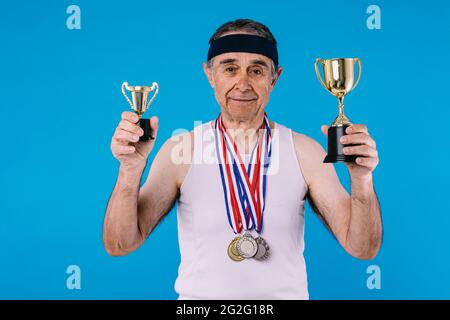 Älterer Athlet mit Sonnenflecken an den Armen, drei Medaillen am Hals und zwei Trophäen in den Händen auf blauem Hintergrund Stockfoto