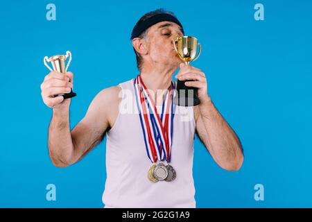 Älterer Athlet mit Sonnenflecken an den Armen, mit drei Medaillen am Hals, die eine Trophäe küssen, auf blauem Hintergrund Stockfoto