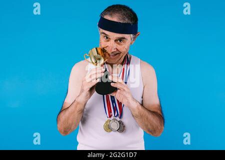 Älterer Athlet mit Sonnenflecken auf den Armen, mit drei Medaillen auf dem Hals, die auf blauem Hintergrund eine Trophäe beißen Stockfoto