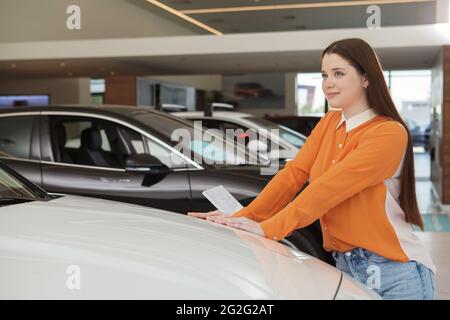 Schöne glückliche junge Frau, die träumerisch wegschaut, während sie sich für ein neues Auto im Autohaus entscheidet Stockfoto