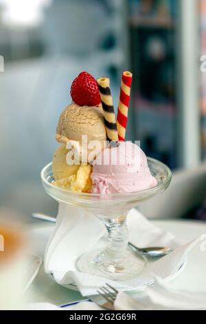 Speiseeis in drei Geschmacksrichtungen, begleitet von bunten Spiralscheiben, die alle in einer schönen Glastasse präsentiert werden, gekrönt mit einer Erdbeere auf der Oberseite. Stockfoto