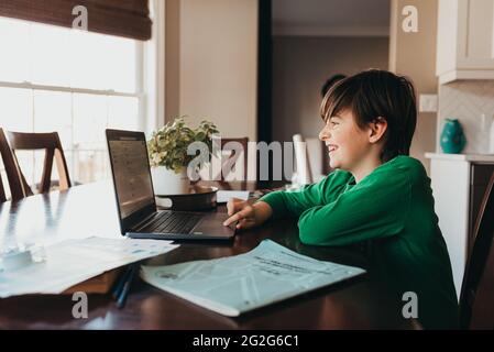 Glücklicher Junge, der auf der Schularbeit online am Computer am Küchentisch arbeitet. Stockfoto