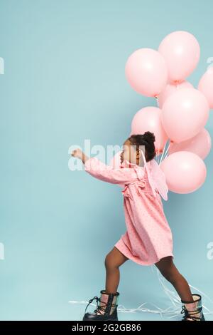 Glückliches schwarzes Mädchen mit Spielzeug Flügel laufen mit rosa Ballons isoliert auf blauem Hintergrund Stockfoto